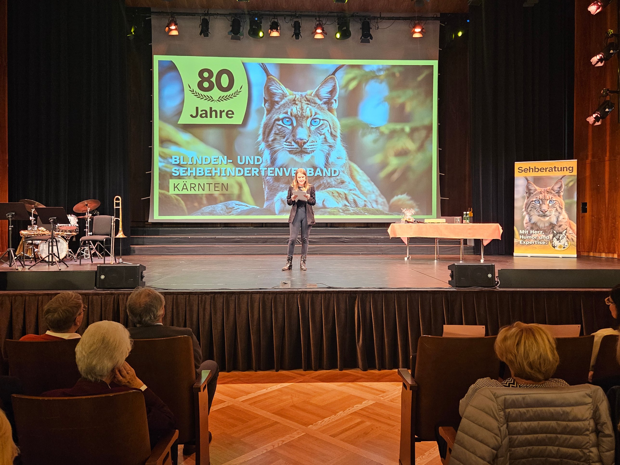 Rednerin bei ihrem Impulsvortrag auf der Bühne des Konzerthauses Klagenfurt vor einer Leinwand mit Luchs und der Aufschrift „80 Jahre Blinden- und Sehbehindertenverband Kärnten“; links die Instrumente der Jazzband, rechts ein Roll-up der Sehberatung, im Vordergrund das Publikum